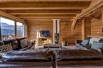Cozy, rustic wooden chalet living room with mountain views through large windows, featuring leather sofas, a wood-burning stove, and TV.