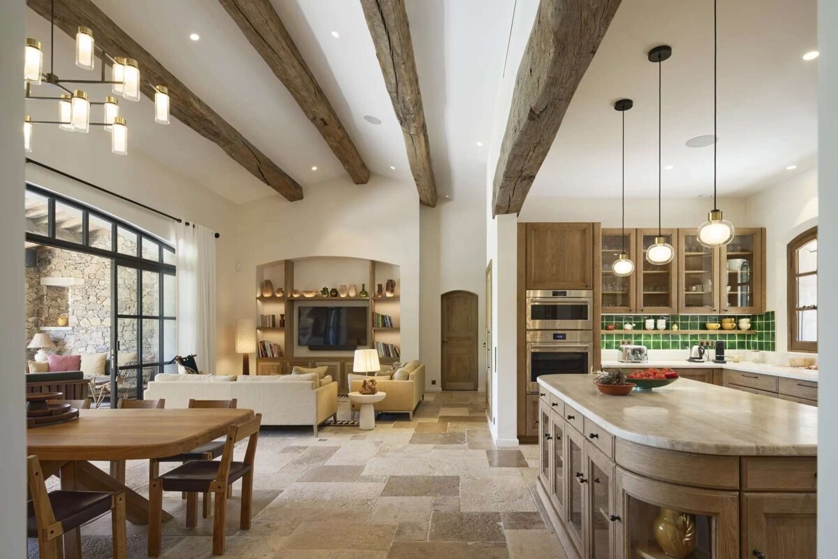 An expansive, rustic open-plan home featuring a dining area, living room, and a modern kitchen with green tile backsplash. Highlights include exposed wooden beams, stone floors, and large windows.