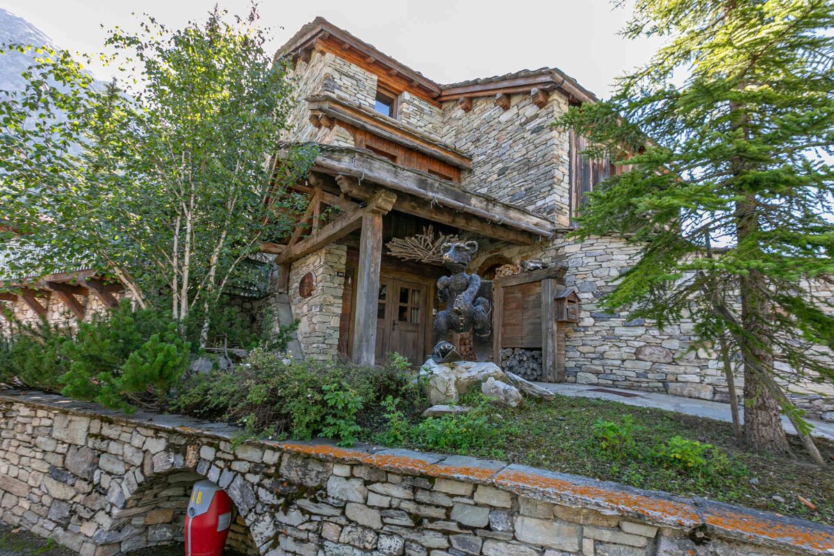 A rustic stone chalet with a prominent wooden bear statue guarding its entrance, surrounded by lush trees and a stone wall.