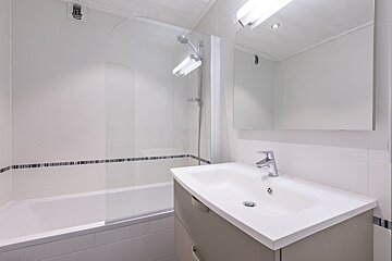 A modern white tiled bathroom with a bathtub, glass shower screen, vanity sink, and mirror with an overhead light.