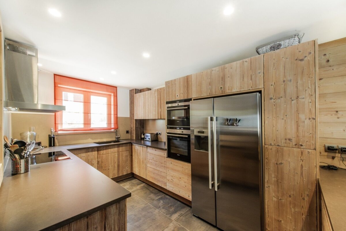 A modern kitchen featuring rustic wooden cabinetry, stainless steel appliances, dark countertops, and a window with a vibrant orange blind.