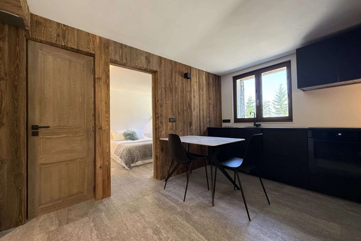 A modern interior with wooden wall panels, a black kitchenette with a dining table, and a bedroom visible through a doorway. A window shows trees outside.