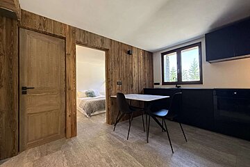 A modern interior with wooden wall panels, a black kitchenette with a dining table, and a bedroom visible through a doorway. A window shows trees outside.