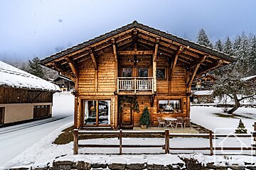 A rustic wooden chalet with a balcony, surrounded by snow in a mountain village. Gentle snowfall creates a picturesque winter scene.