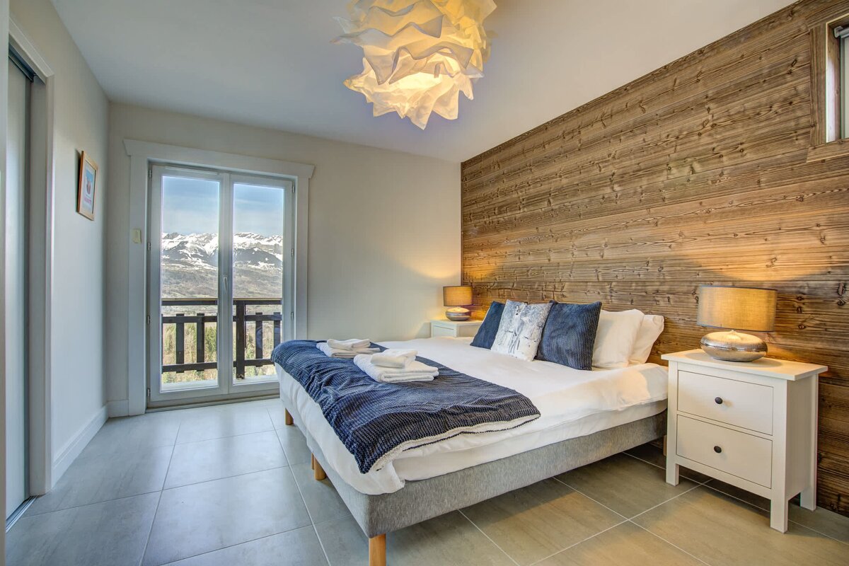 Modern bedroom with a wooden feature wall, cozy bed, and glass balcony doors overlooking snowy mountains.