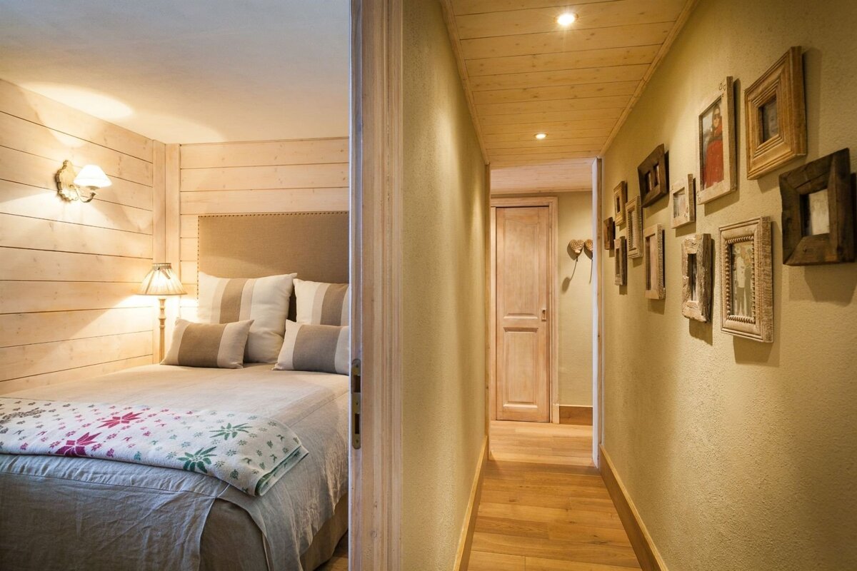 A cozy bedroom with wooden walls and a bed, visible next to a hallway adorned with framed pictures and a wooden floor, all in warm, light tones.