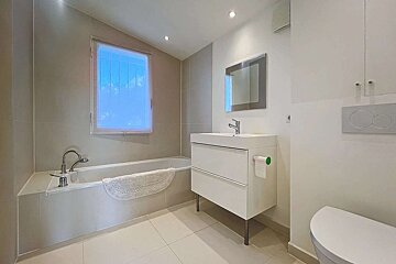 A bright, modern bathroom featuring a white bathtub, vanity with sink, mirror, and toilet. Neutral tiled walls and floor, with a sunlit window.
