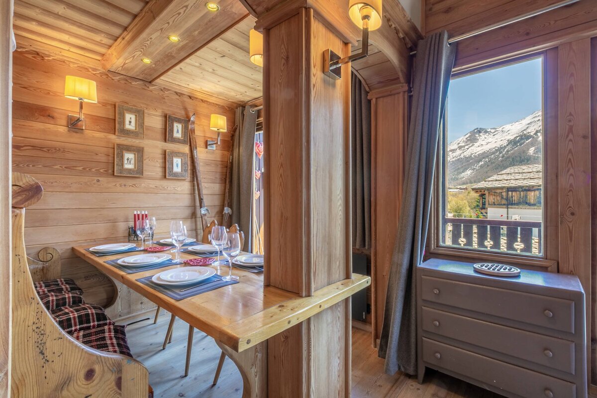 Cozy, wood-paneled chalet dining area with a set table, warm lighting, and a window framing a picturesque view of snow-capped mountains.