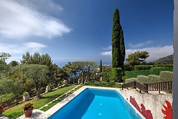 A hillside swimming pool surrounded by lush green trees and shrubs, overlooking a distant sea under a bright blue sky.
