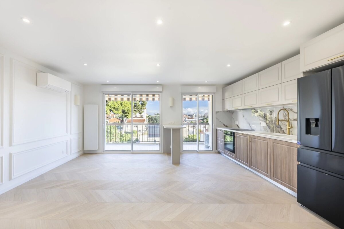 Bright open-plan apartment featuring herringbone floors, a modern kitchen with a black fridge, and sliding doors to a balcony.