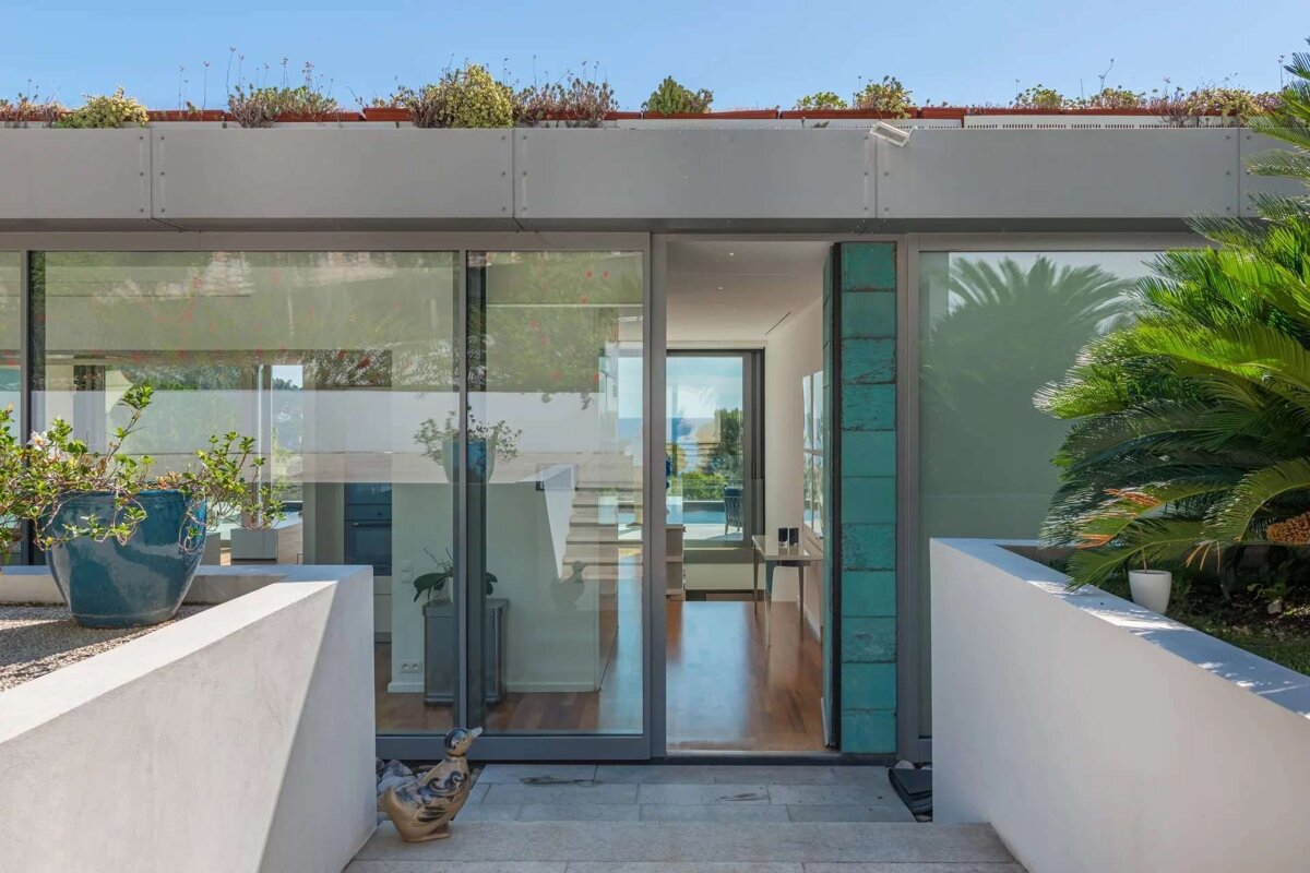 A modern house entrance with large glass doors reveals an interior and distant ocean view. It features a rooftop garden, potted plants, and a duck statue.