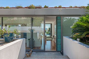 A modern house entrance with large glass doors reveals an interior and distant ocean view. It features a rooftop garden, potted plants, and a duck statue.