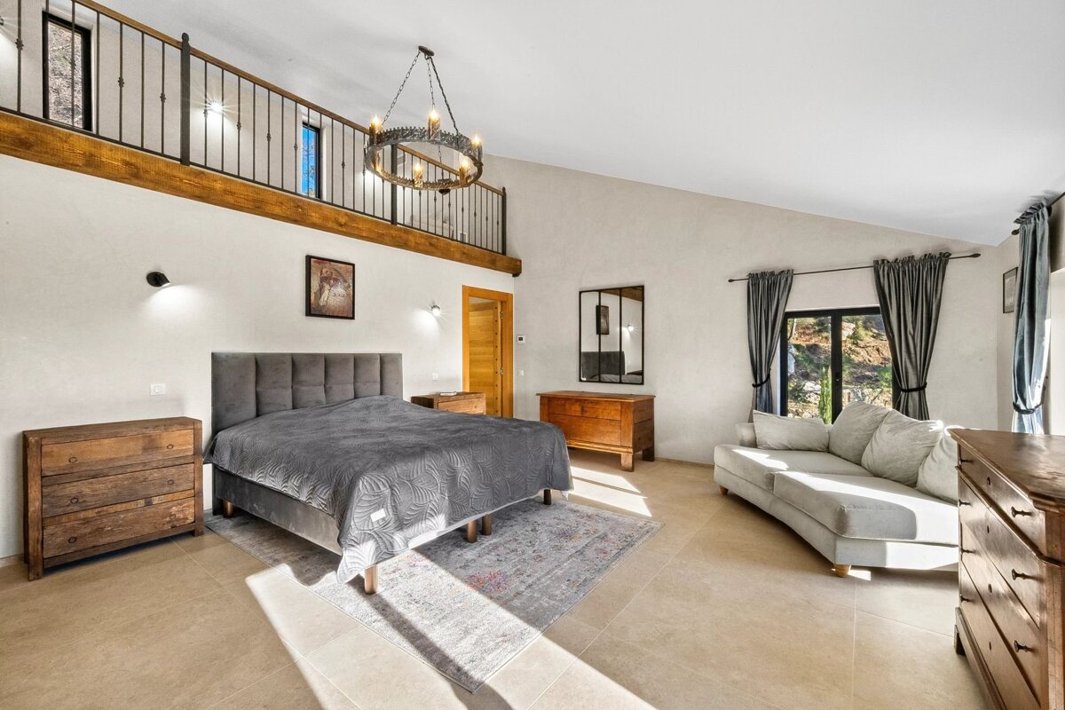 Bright, high-ceilinged bedroom with a loft balcony, large bed, gray sofa, rustic wooden furniture, and an ornate chandelier.