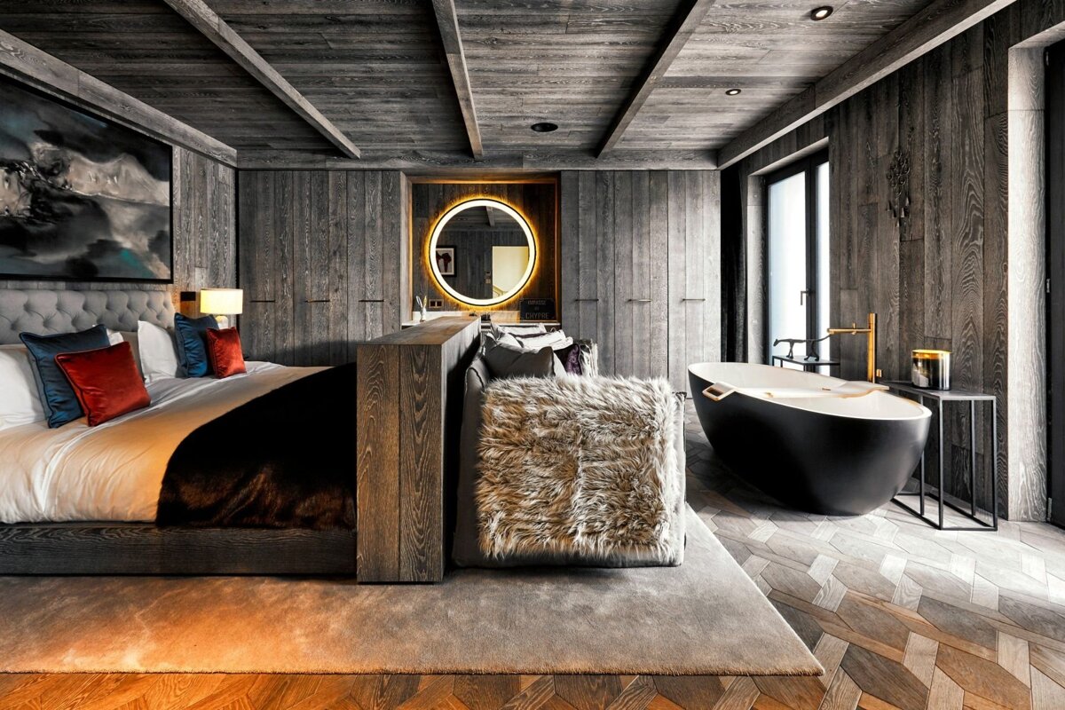 Luxurious modern suite featuring dark wood paneling, a plush bed, a chic black freestanding tub, and an illuminated mirror.