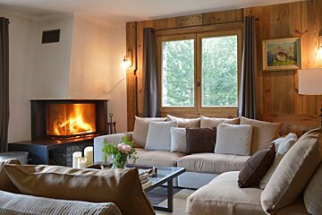 Cozy, rustic living room with a warm fireplace, plush sofas, wood walls, and a window framing a green view.