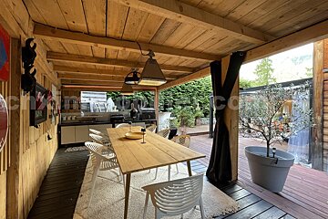 A rustic wooden outdoor patio with a dining area, modern kitchenette, and potted tree. Features a long wooden table, white chairs, and lush green landscaping.