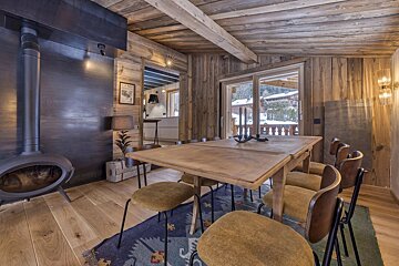 Rustic chalet dining room with a large wooden table, chairs, a black wood-burning stove, and a snowy view through glass doors.