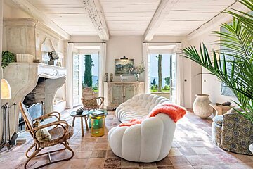 A bright, rustic living room featuring a modern curved white sofa, terracotta floors, exposed beams, and French doors offering serene outdoor views.