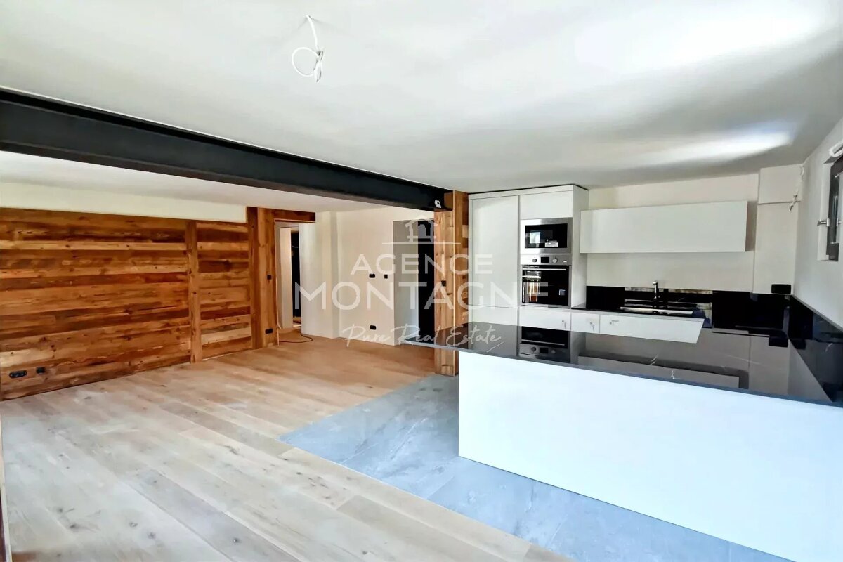 A modern open-plan room featuring a sleek white kitchen with black countertops, a structural beam, and a rustic wood-paneled wall in the living area.