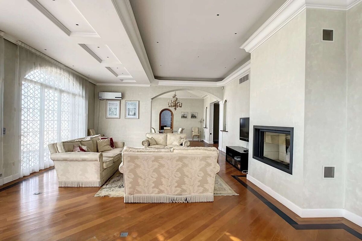 Spacious, elegant living room with ornate sofas, hardwood floors, a modern fireplace, and a large patterned window, leading to a dining area.