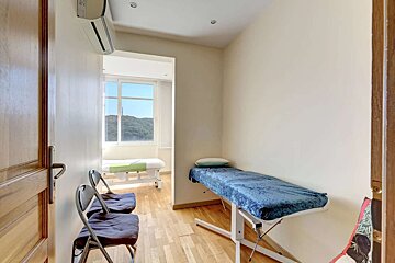 A bright treatment room with two massage tables, two chairs, light walls, and a wooden floor, featuring a window view of green hills.