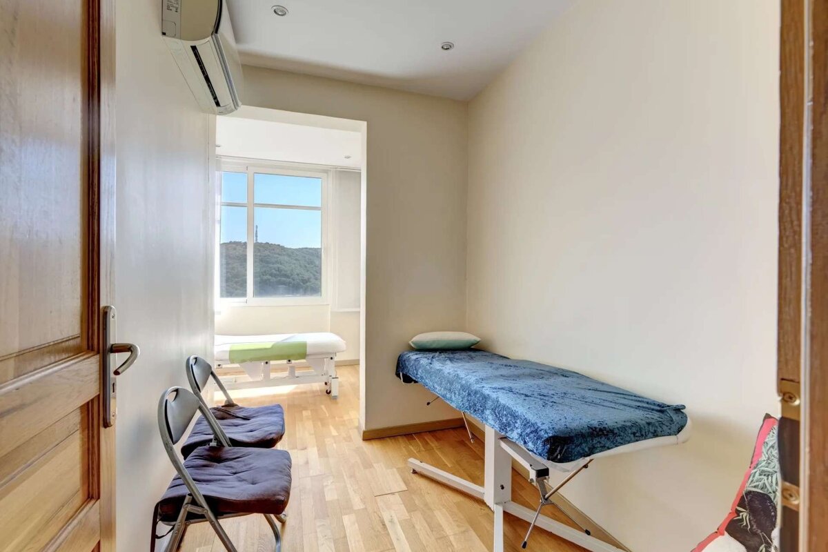 A bright room with two massage/treatment tables, waiting chairs, wooden floor, an AC unit, and a window overlooking green hills.