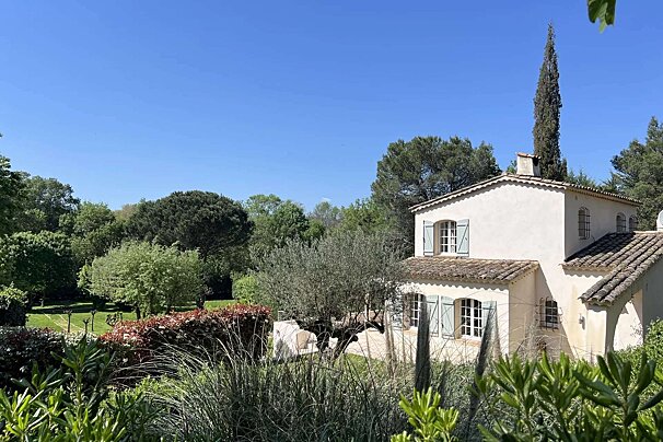 A light-colored house with blue shutters nestled in a lush green garden filled with trees, under a bright clear blue sky.
