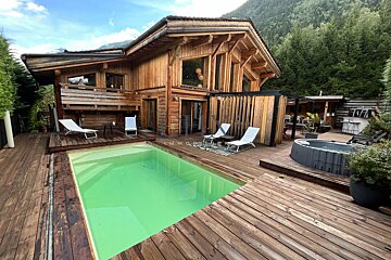 Luxurious wooden chalet with a swimming pool and hot tub on a large deck, surrounded by a mountain forest.