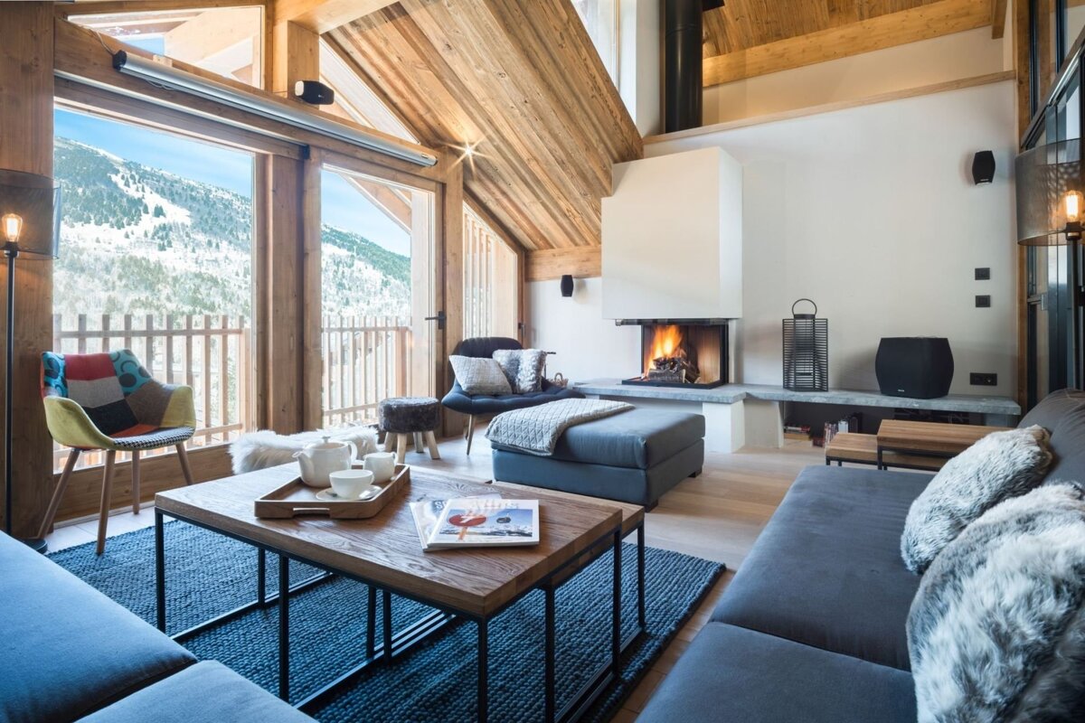 A cozy, modern chalet living room with a fireplace, comfortable seating, and large windows offering a stunning view of snowy mountains.