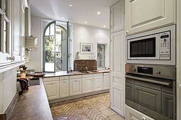 A bright kitchen with white cabinets, brown countertops, and a herringbone tiled floor. An arched green door opens to a lush garden view.