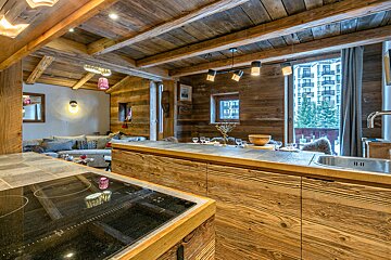 A cozy, rustic alpine chalet's open-plan kitchen and living area. Features rich wood paneling, modern appliances, and a view of buildings and trees.