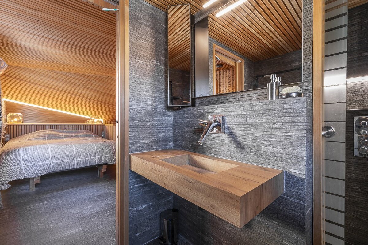 A rustic bathroom with a wooden sink, stone-textured walls, and a view into a cozy bedroom with slanted ceilings.