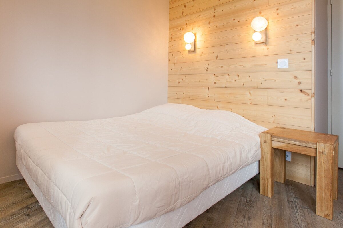 A cozy bedroom featuring a bed with light bedding, a wooden plank headboard, two luminous wall sconces, a small wooden side table, and wooden floors.
