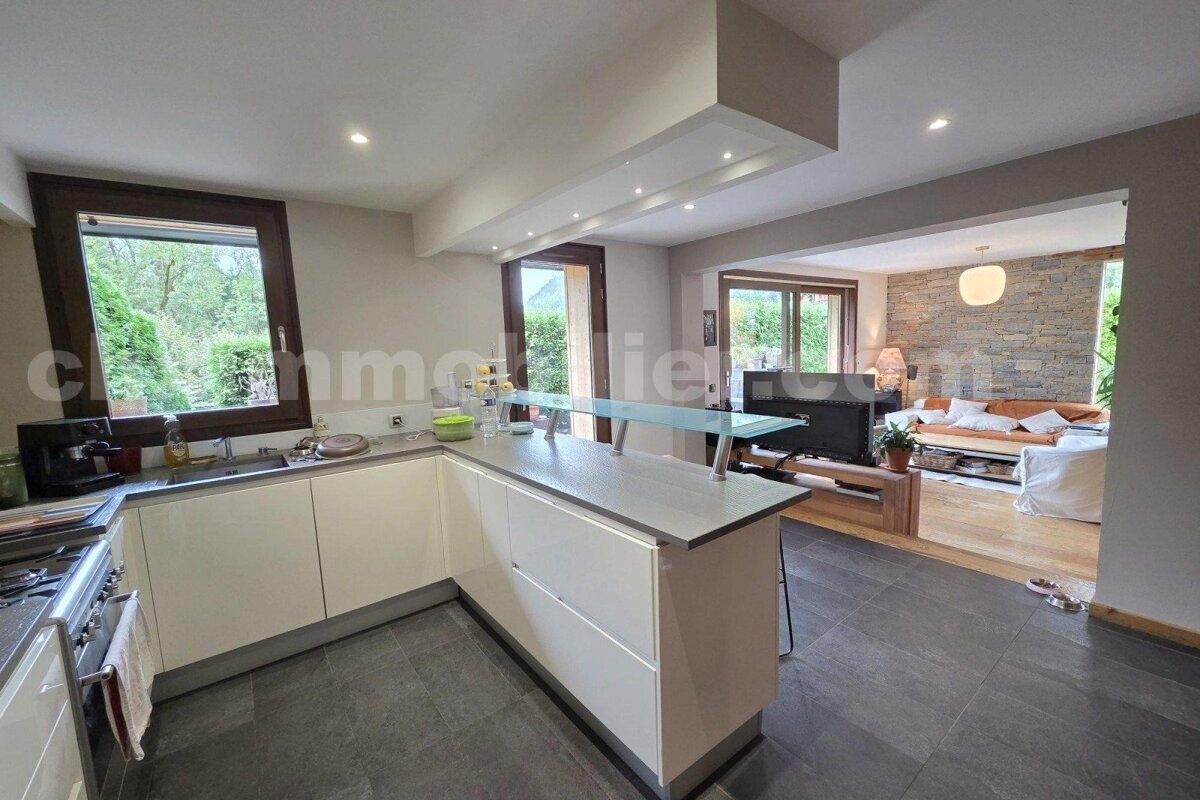A modern open-plan kitchen with white cabinets, dark tiled floor, and a breakfast bar, opening into a living room with a stone wall and wood floor.
