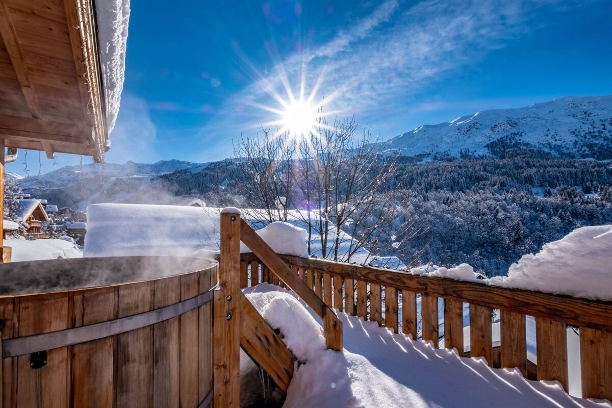 Steaming wooden hot tub on a snowy deck overlooking sunny winter mountains under a brilliant blue sky.