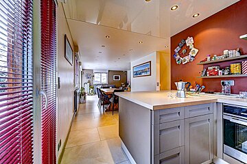 Modern open-plan kitchen and dining area. A kitchen island with a coffeemaker, a red accent wall with shelves, and a long dining table are visible.