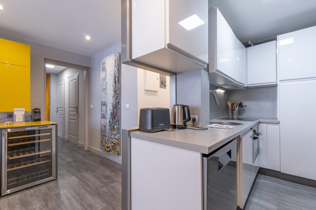 Modern kitchen with sleek white cabinets, grey countertops, a small wine fridge, and a hallway view in the background.