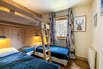 A cozy bedroom features wooden bunk beds and a twin bed, deer-themed curtains, and a window looking out onto snowy trees, creating a warm, cabin-like ambiance.