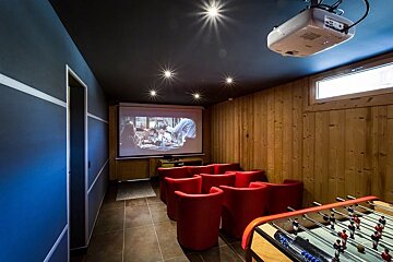 A cozy home cinema room featuring red armchairs, a large projector screen, wood-paneled walls, and a foosball table.