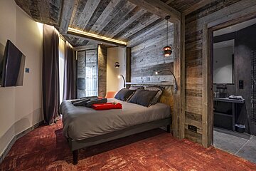 A cozy, rustic bedroom with wood-paneled walls and ceiling, a grey bed on a red carpet, and a view into an adjacent bathroom.