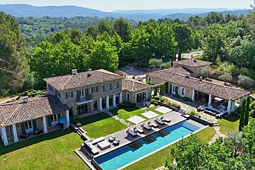 Aerial view of a sprawling luxury estate with multiple stone buildings, a large swimming pool, and lush green lawns, surrounded by forests and hills.