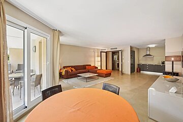 Modern open-plan apartment with dining area, brown living room sofa, sleek kitchen, and balcony access. Orange accents throughout.