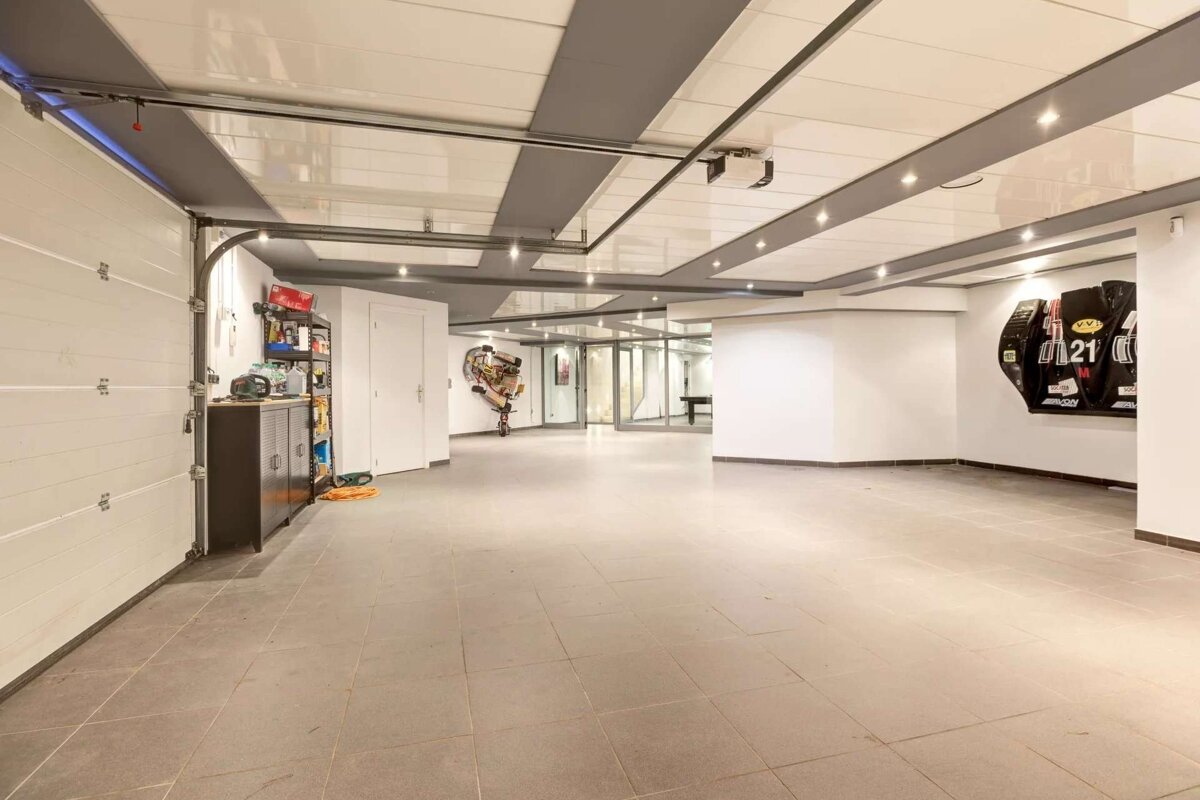 A spacious, modern garage or basement with a roll-up door, tiled floor, bright strip lighting, some storage, and contemporary art on the walls.