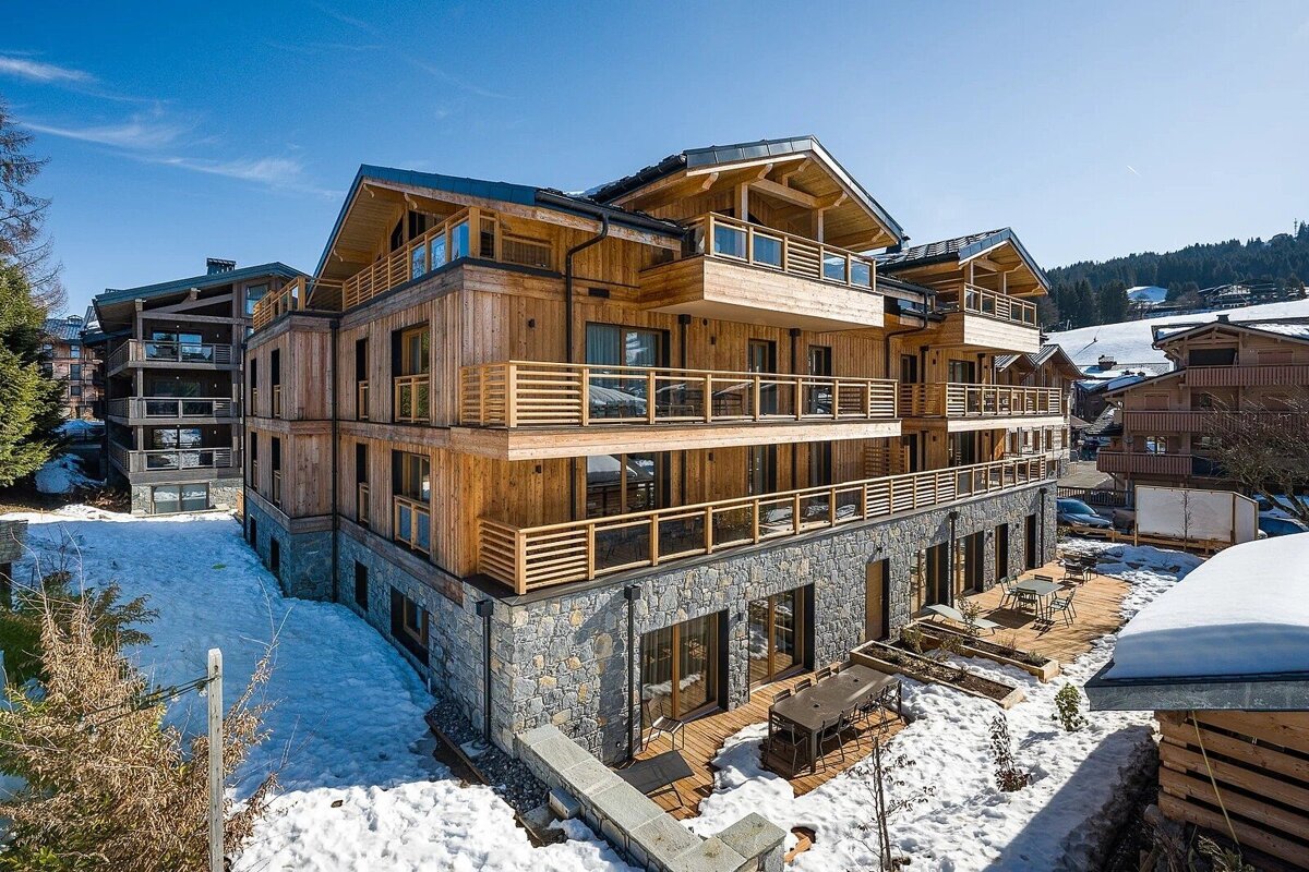Modern multi-story wooden and stone chalet with balconies and outdoor decks, surrounded by snow under a bright blue sky.