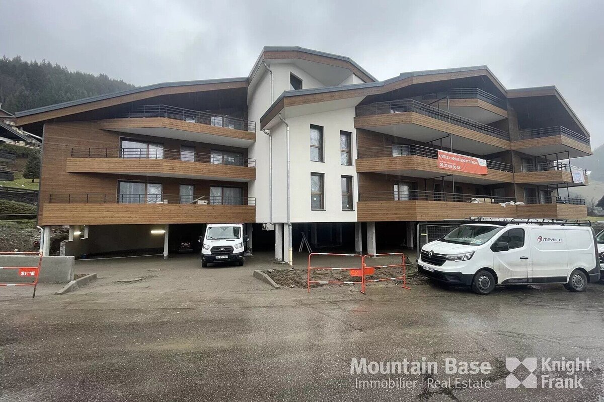 A modern, multi-story building with wood and white facade, elevated over parking, with two white vans in front and a cloudy mountain backdrop.