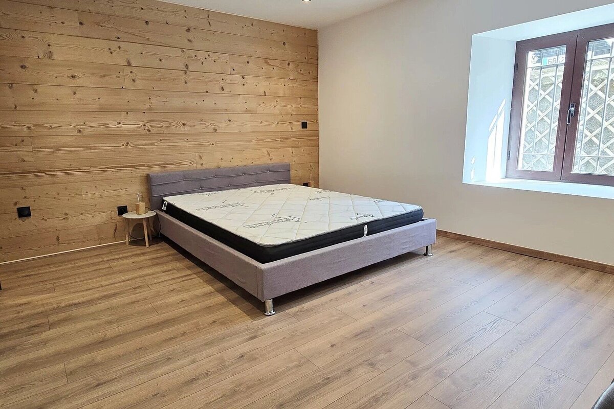 A bedroom with a wooden plank accent wall, grey upholstered bed, light wood floor, and a window.
