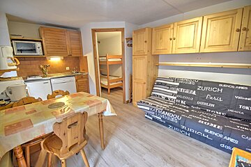 Cozy wood-accented studio apartment with a dining table, kitchen, a printed futon sofa, and bunk beds visible in an adjoining room.