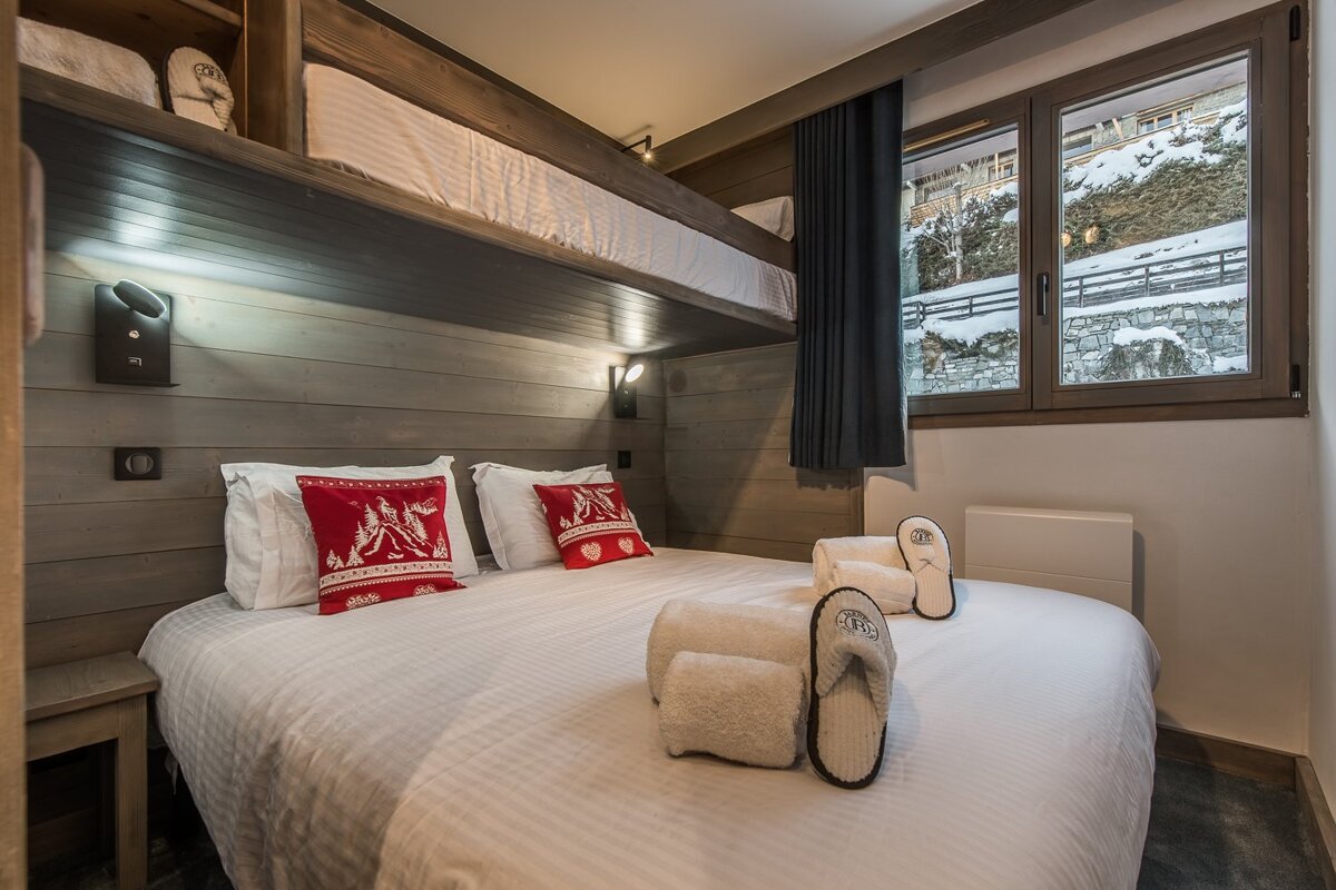 Cozy bedroom with a double bed, red pillows, and a bunk bed above, featuring wooden walls and a window overlooking a snowy landscape.