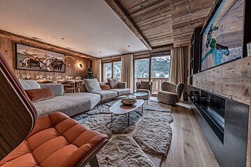 A warm, wood-paneled ski chalet living room and dining area, featuring plush seating, a large shaggy rug, and expansive windows with snowy mountain views.