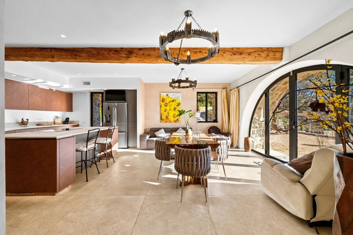 A bright open-plan kitchen, dining, and living space with rustic wood beams, modern fixtures, and a large arched window overlooking nature.
